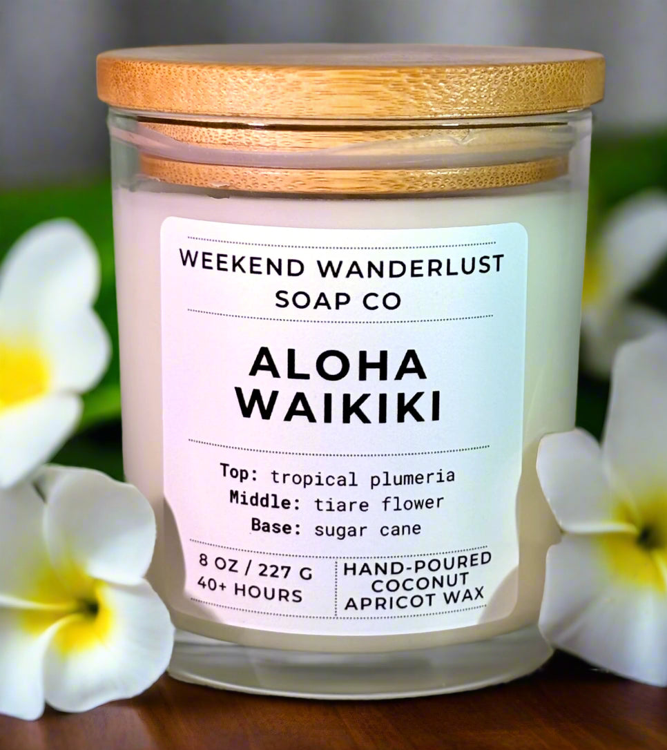 Candle with 'Weekend Wanderlust Soap Co' label and 'Aloha Waikiki' description, surrounded by flowers.