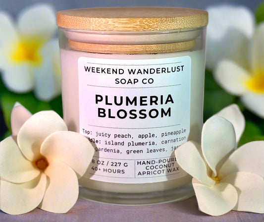 Candle labeled 'Plumeria Blossom' from Weekend Wanderlust Soap Co with plumeria flowers in the background.