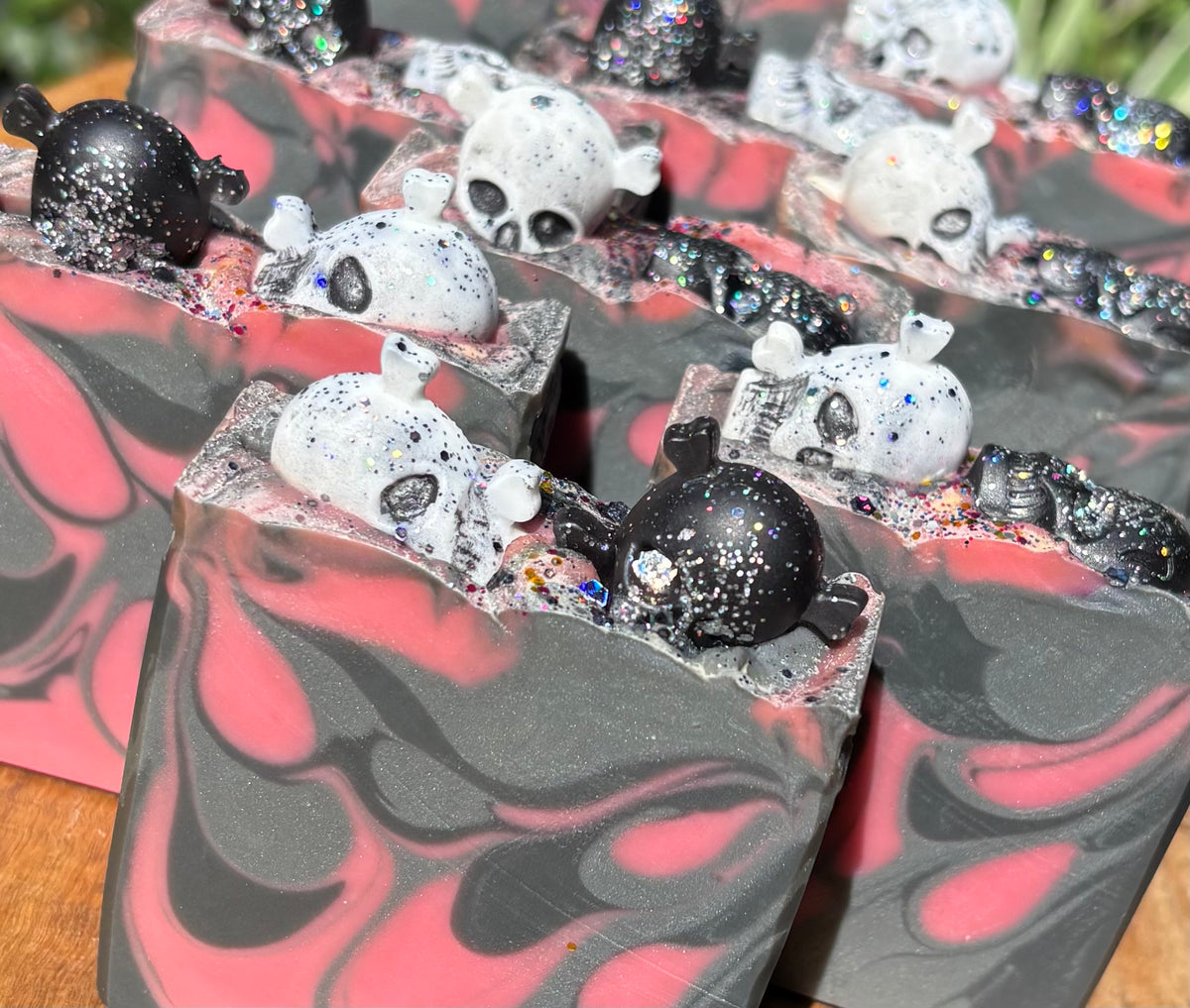 A close-up of several handmade rectangular bars of soap with a marbled black and pink pattern, topped with small white and black skull embellishments and glitter, arranged on a wooden surface.