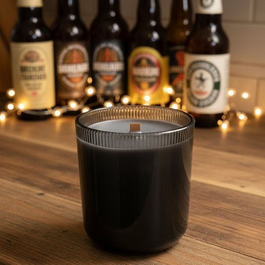 Black candle in a glass container on a wooden surface with beer bottles in the background.