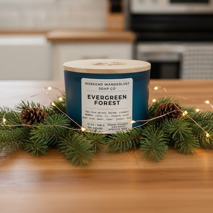 Candle labeled 'Evergreen Forest' from 'Weekend Wanderlust Soap Co.' on a kitchen counter with greenery and lights.