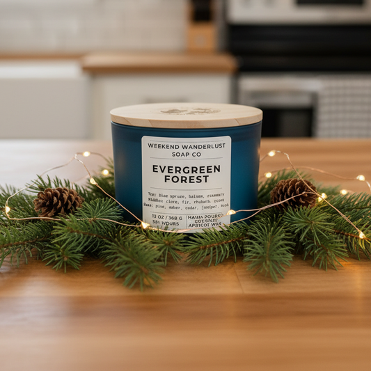 Candle labeled 'Evergreen Forest' from 'Weekend Wanderlust Soap Co.' on a kitchen counter with greenery and lights.