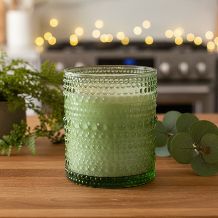 Green textured glass candle holder on a wooden surface with blurred lights in the background